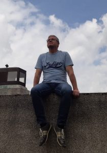 Frits Stiemer, kunstenaar, zittend op een muur met een Indigo t-shirt aan